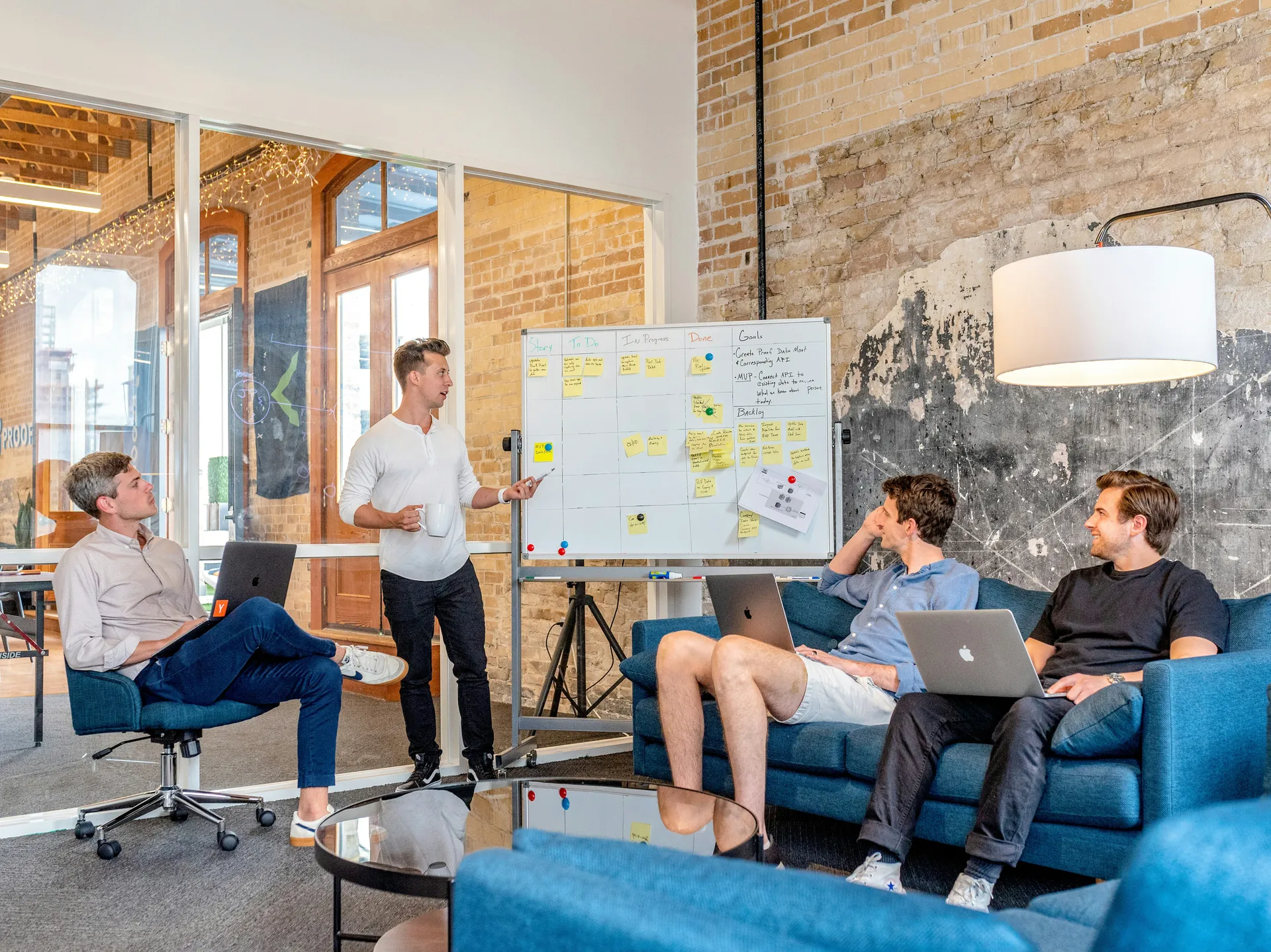 Réunion de quatre hommes dans un bureau moderne discutant devant un tableau blanc couvert de post-its, avec des ordinateurs portables.