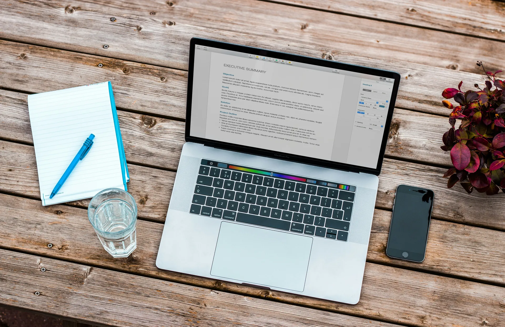 Ordinateur portable affichant un document sur une table en bois, avec un carnet, un stylo, un verre d'eau et un smartphone à côté