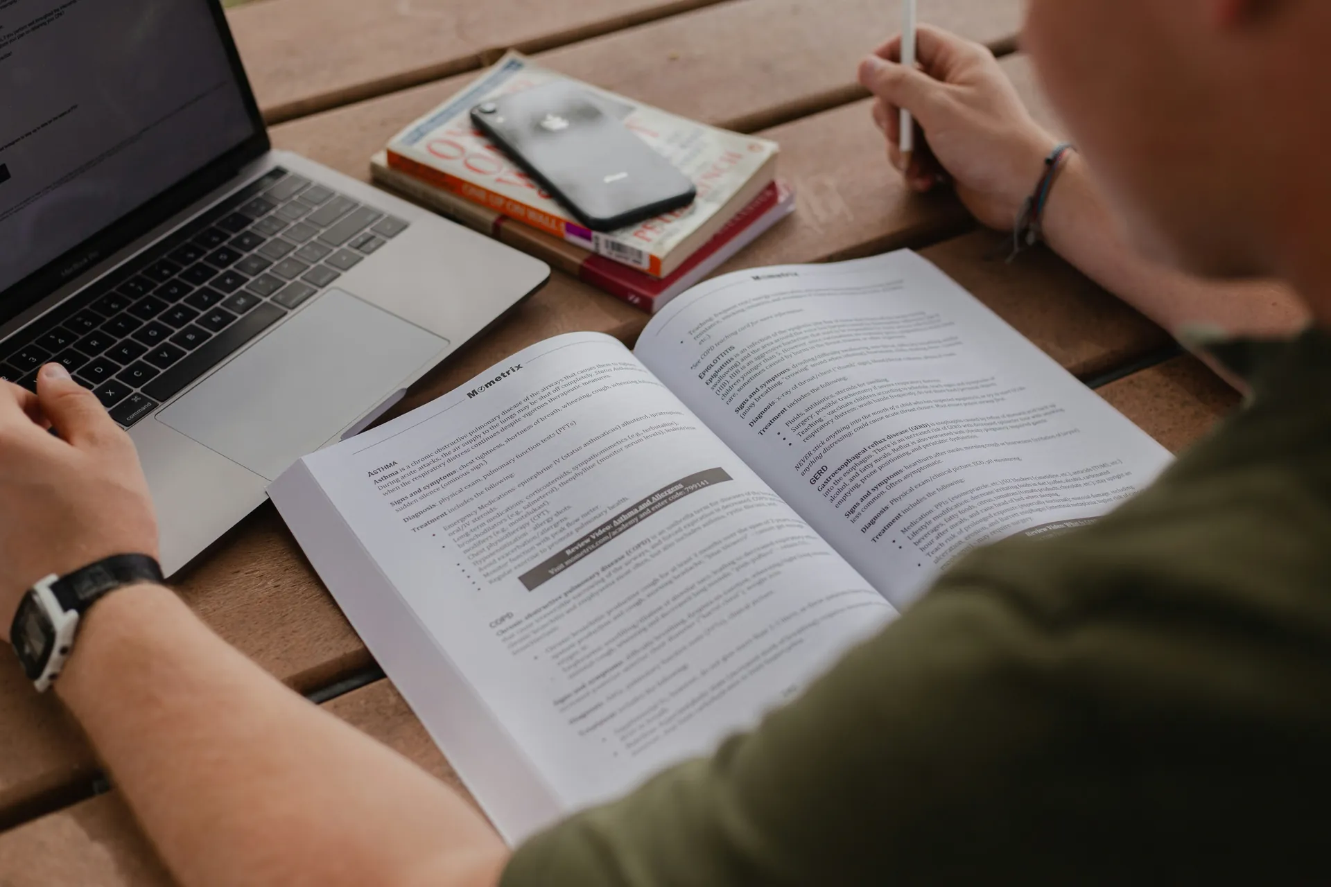 Student studying an open exam prep book beside a laptop for certification exam prep