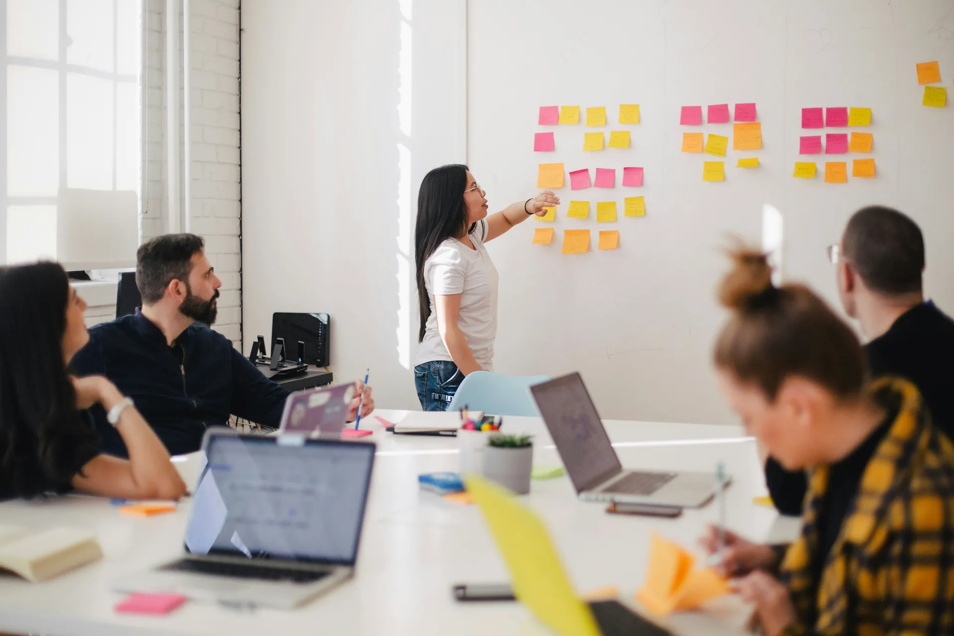 Réunion d'équipe en salle de travail, une femme debout présente un tableau rempli de post-its colorés pendant que ses collègues prennent des notes et écoutent autour d'une table.