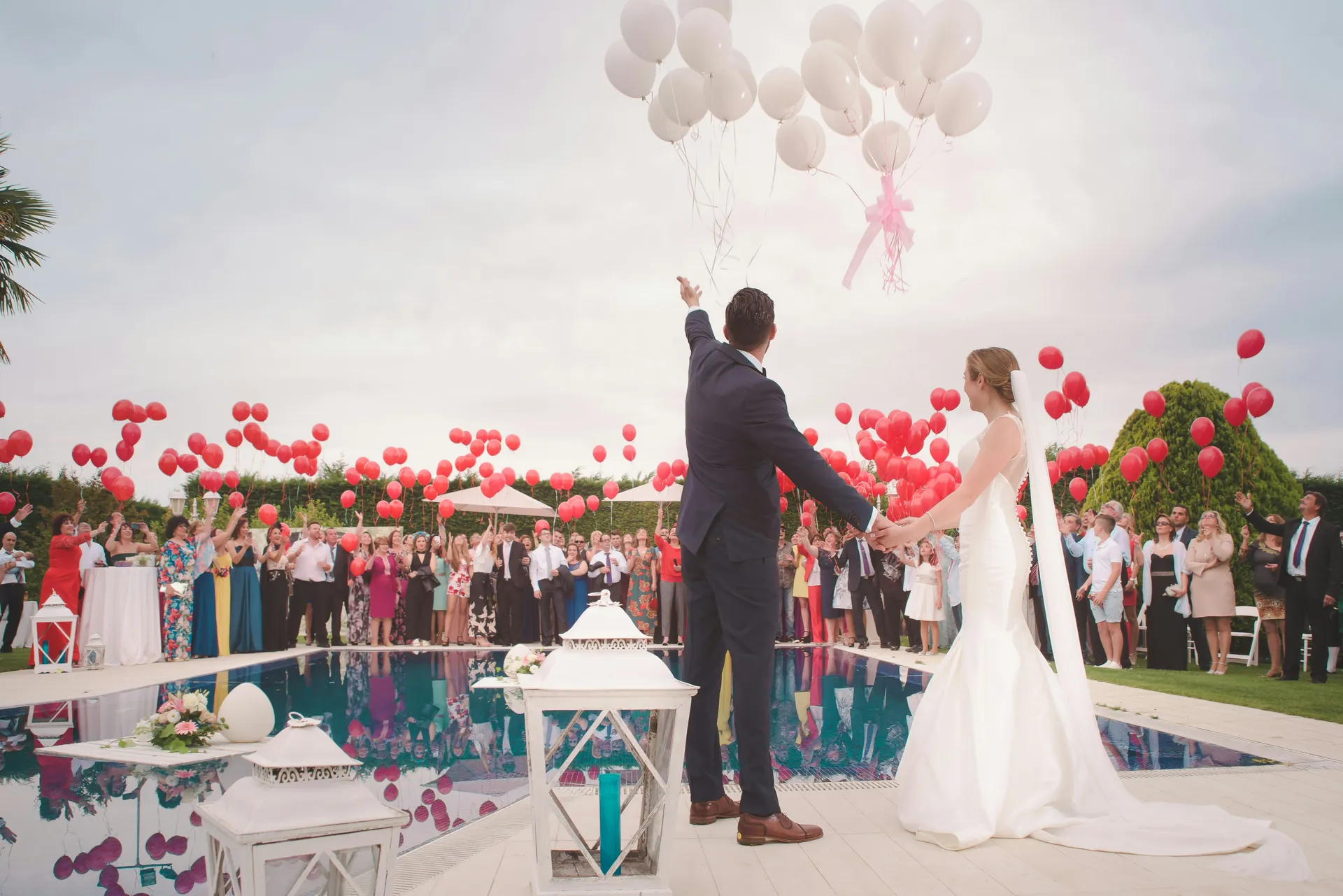 Couple getting married by a poolside in the Bahamas, surrounded by guests releasing red and white balloons, celebrating their wedding in a tropical outdoor setting.